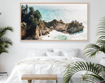 May include: A framed photograph of a waterfall cascading down a rocky cliff into a turquoise ocean. Lush green trees and foliage surround the waterfall. The photo is displayed on a white wall above a white bed with a white blanket and a wooden bench.