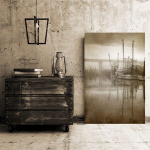 May include: A sepia-toned photograph of a fishing boat docked in a harbor, with a bridge in the background. The boat is surrounded by fog and the water is calm and reflective. The photograph is printed on canvas and mounted on a wooden frame.