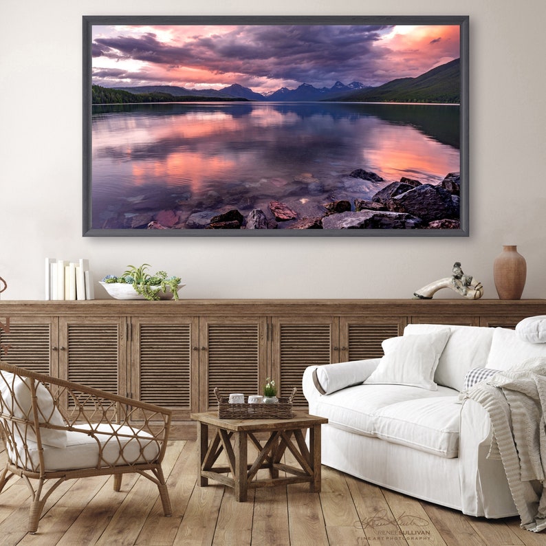 Pu&ograve; includere: Una fotografia panoramica di un lago con montagne sullo sfondo. Il cielo &egrave; un mix vibrante di sfumature rosa, viola e arancioni, che si riflettono nell'acqua calma. Il primo piano presenta una costa rocciosa.