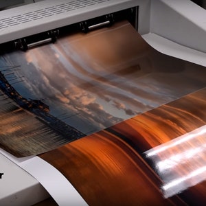 Puede incluir: Primer plano de una impresora grande con dos hojas de papel met&aacute;lico que se introducen en la m&aacute;quina. El papel tiene una imagen de puesta de sol impresa. El texto "Metallic Paper" es visible en la esquina inferior izquierda de la imagen.