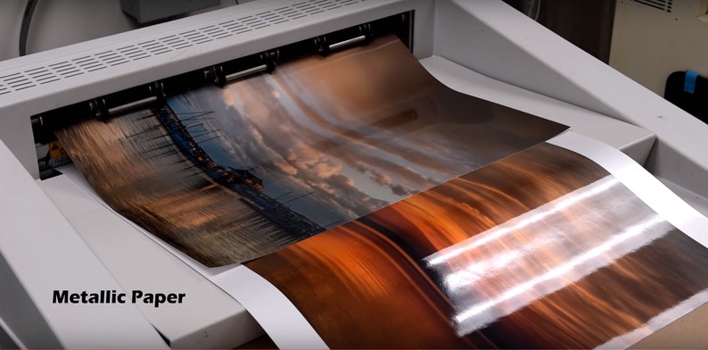 Puede incluir: Primer plano de una impresora grande con dos hojas de papel met&aacute;lico que se introducen en la m&aacute;quina. El papel tiene una imagen de puesta de sol impresa. El texto "Metallic Paper" es visible en la esquina inferior izquierda de la imagen.