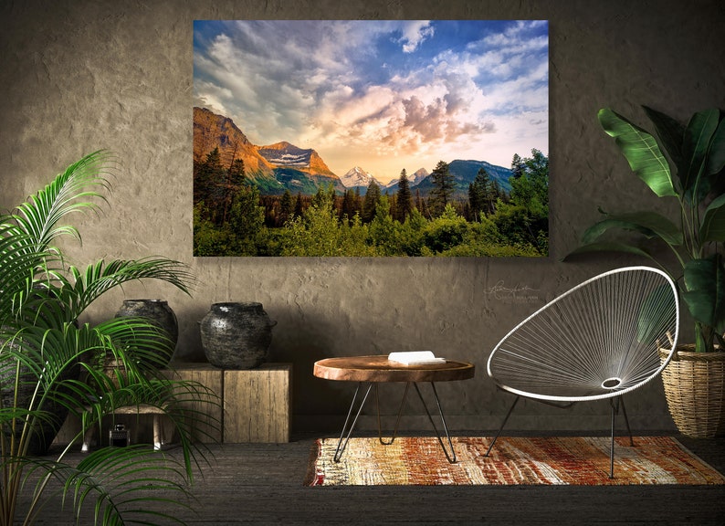 Op de afbeelding: Een woonkamer interieur met een grote ingelijste landschapsfoto van een bergketen met een bewolkte lucht. De foto hangt aan een getextureerde muur. Er is een houten salontafel met een boek erop, een witte draadstoel en twee planten in potten.