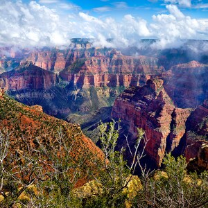 May include: A panoramic view of the Grand Canyon with a dramatic rock formation in the foreground. The canyon is filled with layers of red rock and is partially obscured by clouds.
