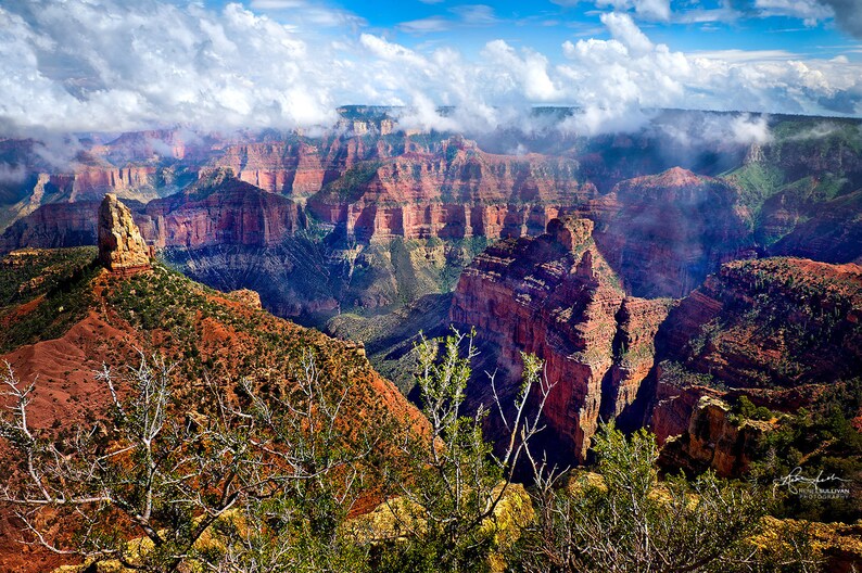 May include: A panoramic view of the Grand Canyon with a dramatic rock formation in the foreground. The canyon is filled with layers of red rock and is partially obscured by clouds.