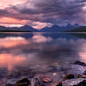 Pu&ograve; includere: Una vista panoramica di un lago con montagne sullo sfondo. Il cielo &egrave; di un rosa e viola vibrante, che si riflette nell'acqua calma. In primo piano, le rocce costeggiano la riva.