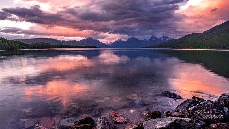 Pu&ograve; includere: Una vista panoramica di un lago con montagne sullo sfondo. Il cielo &egrave; di un rosa e viola vibrante, che si riflette nell'acqua calma. In primo piano, le rocce costeggiano la riva.