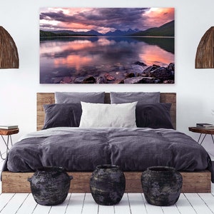 Pu&ograve; includere: Un design d'interni per una camera da letto con una grande stampa su tela di un lago e montagne al tramonto. La stampa &egrave; appesa sopra un letto con un piumone grigio scuro e una testiera in legno. Ci sono due comodini in legno con lampade e una piccola pianta in vaso.