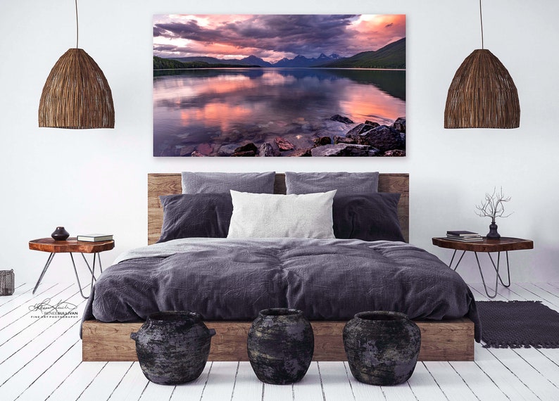 Pu&ograve; includere: Un design d'interni per una camera da letto con una grande stampa su tela di un lago e montagne al tramonto. La stampa &egrave; appesa sopra un letto con un piumone grigio scuro e una testiera in legno. Ci sono due comodini in legno con lampade e una piccola pianta in vaso.