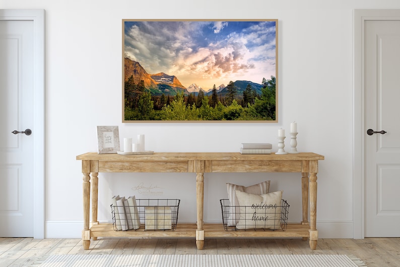 Op de afbeelding: Een ingelijste landschapsfoto van een bergketen met een bewolkte lucht en zonsondergang. De foto hangt aan een witte muur boven een houten consoletafel met twee manden en kaarsen.