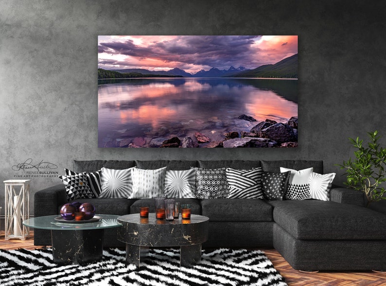 Pu&ograve; includere: Una fotografia panoramica di un lago con montagne sullo sfondo. Il cielo &egrave; di un rosa e viola vibrante con nuvole. Il lago &egrave; calmo e riflette i colori del cielo. Ci sono rocce in primo piano.