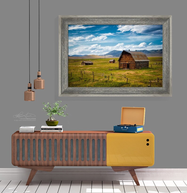 Puede incluir: Una fotograf&iacute;a enmarcada de un granero de madera r&uacute;stico en un campo con un cielo azul y nubes blancas. El granero est&aacute; rodeado de hierba verde y una valla de madera. La foto est&aacute; colgada en una pared gris sobre un tocadiscos vintage y un armario de madera.