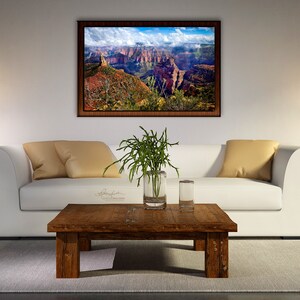 May include: A white couch with two beige pillows sits in front of a framed landscape photograph of a canyon. A wooden coffee table with a vase of green plants and two glasses sits in front of the couch. Two lamps with white shades and brown bases are on end tables on either side of the couch.