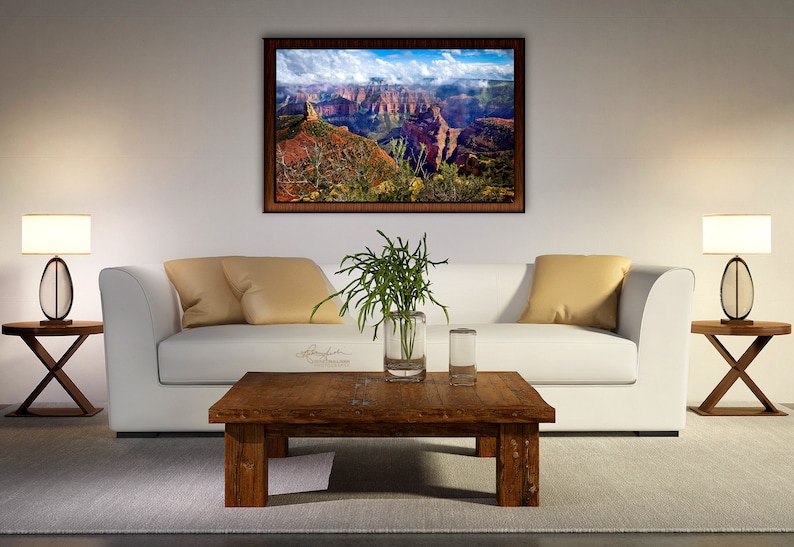 May include: A white couch with two beige pillows sits in front of a framed landscape photograph of a canyon. A wooden coffee table with a vase of green plants and two glasses sits in front of the couch. Two lamps with white shades and brown bases are on end tables on either side of the couch.