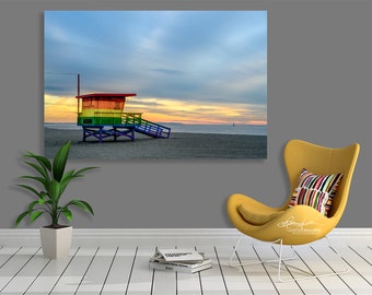 May include: A colourful lifeguard stand painted in rainbow colours stands on a sandy beach with a cloudy sky and a sunset in the background.