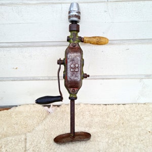 May include: A vintage green and brown hand drill with a wooden handle and a metal chuck. The drill has a crank handle and a metal base.