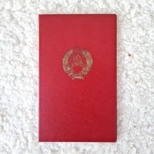 May include: A red book with a gold emblem in the center. The emblem features a wreath surrounding a hammer and sickle, with text below. The book has a textured surface.