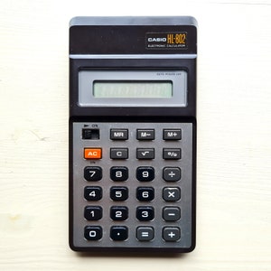 May include: A black Casio HL-802 electronic calculator with a silver face and orange buttons. The calculator has a display screen, a power button, and buttons for numbers, operations, and functions.
