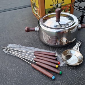 Può includere: Un pentolino per fondue in argento con un manico e un coperchio in legno. Il pentolino è circondato da sei forchette per fondue con manici in legno e punte colorate. C'è anche un piccolo piatto in argento con un manico.