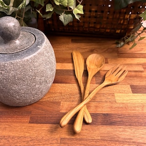 May include: Three wooden utensils, a fork, a spoon, and a knife, are arranged on a wooden surface. The utensils are made of light brown wood and have a natural finish.