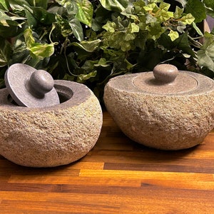 May include: Two gray stone mortar and pestle sets with lids. The mortar and pestle are made of rough, textured stone. The lids are slightly domed and have a small knob on top.