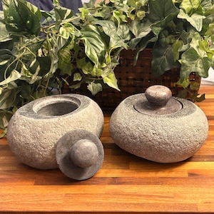 May include: Two gray stone containers with lids. The containers are shaped like rounded rocks. The lids are also made of stone and have a smooth, rounded top.