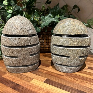 May include: Two stone egg-shaped decorative objects with horizontal cut-out lines. The objects are light gray with a textured surface. The top half of each egg is removable. The objects are on a wooden surface.