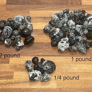 May include: Three piles of dark, round objects with white patches are displayed on a wooden surface. The piles are labeled "1/2 pound", "1 pound", and "1/4 pound". The objects appear to be a type of food or mineral.