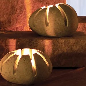 May include: Two stone candle holders with a carved design. The candle holders are oval-shaped and have vertical cuts that allow light to shine through. The stone is a light brown color, and the light inside the candle holders is a warm yellow.