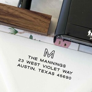 May include: A white envelope with a black address stamp. The stamp reads "M" above "THE MANNINGS, 23 WEST VIOLET WAY, AUSTIN, TEXAS 45690". A wooden stamp handle and a black stamp are also visible.