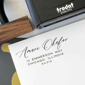 May include: A black Trodat printy 4914 stamp with a wooden handle is next to a white envelope. The envelope has the name "Amari Okafor" in elegant script, along with an address: 12 Emmerson Way, Chicago, Illinois 54312.