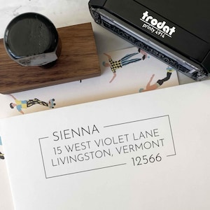 May include: A black address stamp and a wooden stamp handle sit near a white envelope with the name "SIENNA" and address details in black text. The stamp reads "trodat printy 4914".