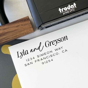 May include: A black Trodat printy 4914 stamp rests on a white envelope. The envelope is printed with the names "Lyla and Greyson" in a cursive font, along with their address: 1234 Simeon Way, San Francisco, CA 91254. A wooden stamp handle is visible.