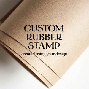 May include: A stack of light brown paper with the words "CUSTOM RUBBER STAMP" in black, with the text "created using your design" below. The paper has a slightly textured appearance and is angled towards the bottom right.