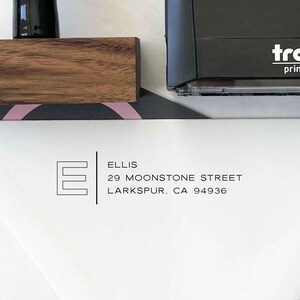 May include: A black address stamp with the text "tro print" is next to a wooden block and a white paper with the address "ELLIS, 29 MOONSTONE STREET, LARKSPUR, CA 94936" and a stylized letter "E".