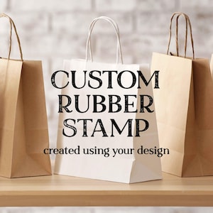 May include: Three shopping bags, two brown and one white, are arranged on a wooden surface. The white bag prominently displays the text "CUSTOM RUBBER STAMP" and "created using your design" in black lettering.