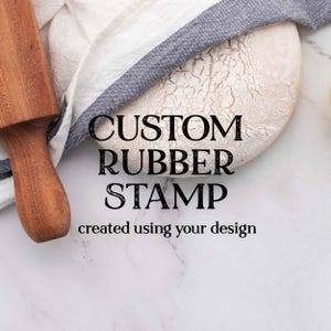 May include: A wooden rolling pin rests next to a loaf of bread and a white and blue striped towel. The text "CUSTOM RUBBER STAMP created using your design" is displayed in black letters.
