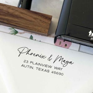 May include: A white envelope with the names "Phoenix & Maya" in elegant script, along with an address. A wooden stamp handle and a black stamp are visible. The address reads: 23 Plainview Way, Autin, Texas 45690.