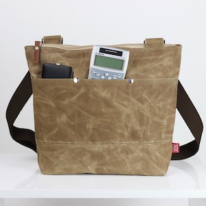 May include: A tan canvas tote bag with a brown shoulder strap. The bag has a zippered top and an exterior pocket with a black wallet and a calculator. A red tag is attached to the side.