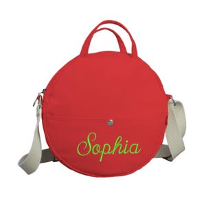 May include: Red round canvas bag with a white strap and a green embroidered name "Sophia".