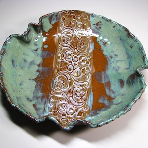May include: A round ceramic bowl with a ruffled, scalloped edge. The bowl features a central vertical band with an embossed swirling vine pattern in a deep brown glaze, flanked by mottled light green and brown glazed sections on both sides.