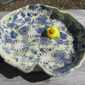 May include: A decorative ceramic bowl with a floral pattern in blue and yellow. The bowl contains a yellow rubber duck and several white flower-shaped decorations. The bowl is on a wooden surface.