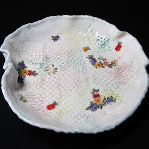 May include: White ceramic serving dish with a scalloped edge. The dish is decorated with a delicate net pattern and colorful floral accents in red, yellow, blue, and green. A unique piece for serving.