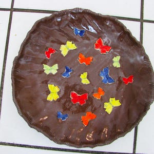 May include: A brown ceramic plate with a textured rim, decorated with colorful butterfly designs. The butterflies are in shades of red, yellow, blue, and orange. The plate is on a white tiled surface.