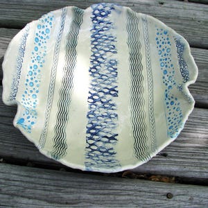 May include: A handmade ceramic serving dish with a scalloped edge. The dish features a cream-colored base with vertical stripes in varying patterns of blue. The patterns include wavy lines, dotted lines, and fish scale designs. The dish is round and appears to be hand-painted.