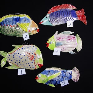 May include: Five ceramic fish wall decorations labeled 1 through 5. Fish 1 is light green with blue dots. Fish 2 is yellow, blue, and green with red accents. Fish 3 is blue with red fins. Fish 4 is light green and pink. Fish 5 is blue and green.