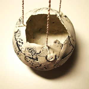 May include: A handmade, spherical ceramic planter with a textured, off-white surface and black decorative patterns. The planter is suspended by a woven cord. The top of the planter is open, revealing the interior.