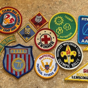 May include: A collection of colorful embroidered patches, including Cub Scout, Girl Scout, and Olympic Rally designs. The patches feature various shapes, text, and imagery, such as a diamond, a sun, and a cross. Some patches have text like "Fitness USA" and "Swimmer".