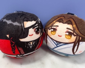 Può includere: Due peluche rotonde con volti in stile anime. Il giocattolo a sinistra è rosso con capelli neri e una benda nera sull'occhio. Il giocattolo a destra è bianco con capelli castani e occhi gialli.