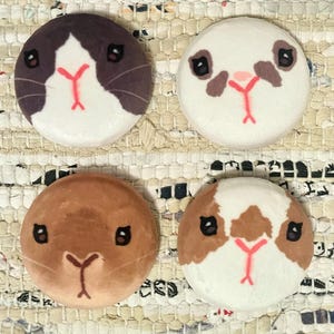 Pode incluir: Quatro bombas de banho redondas e brancas com rostos de animais pintados. Os rostos são um porquinho-da-índia marrom e branco, um porquinho-da-índia marrom, um porquinho-da-índia preto e branco e um porquinho-da-índia branco com detalhes marrons.