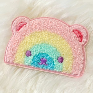 Op de afbeelding: Een roze pluche regenboog patch met een berenkop in het midden. De regenboog is geel, groen, blauw en paars. De berenkop heeft paarse ogen en een neus.
